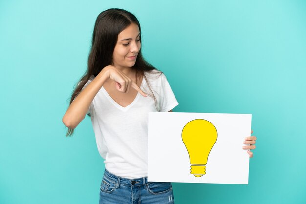 Foto giovane donna francese isolata su priorità bassa blu che tiene un cartello con l'icona della lampadina e lo indica