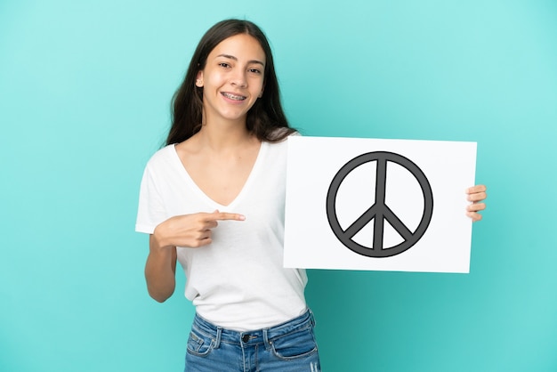 Young French woman isolated on blue background holding a placard with peace symbol and  pointing it