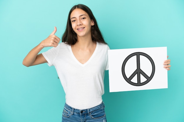 Young French woman isolated on blue background holding a placard with peace symbol with thumb up