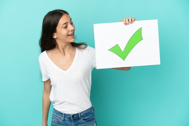 Young French woman isolated on blue background holding a placard with text Green check mark icon with happy expression