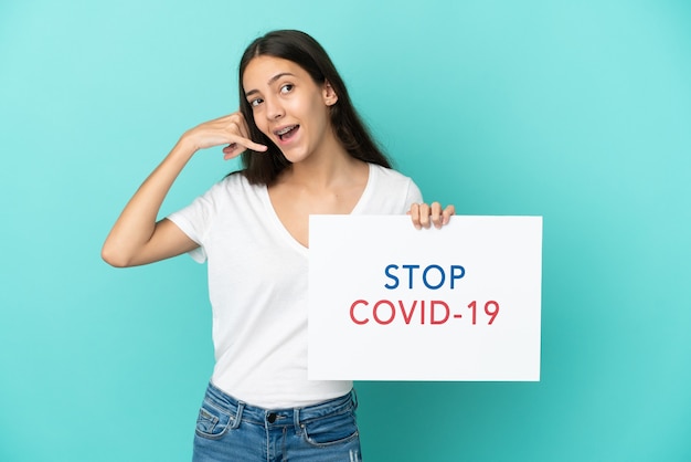 Young French woman isolated on blue background holding a placard with text Stop Covid 19 and doing phone gesture