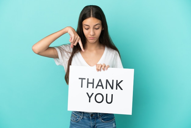 Young French woman isolated on blue background holding a placard with text THANK YOU and  pointing it