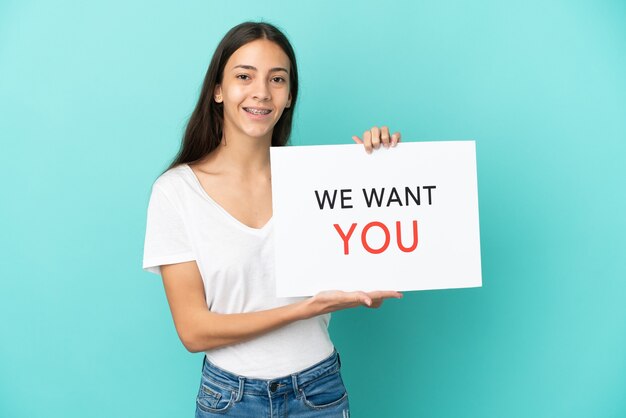 Young French woman isolated on blue background holding We Want You board with happy expression