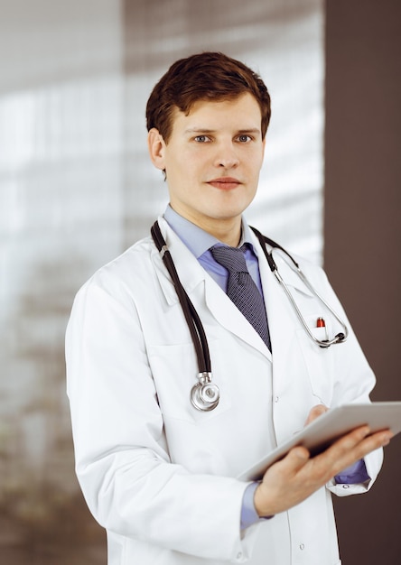 Young friendly doctor is checking some information on his computer tablet. Portrait of a professional physician at work in a clinic. Medicine concept.