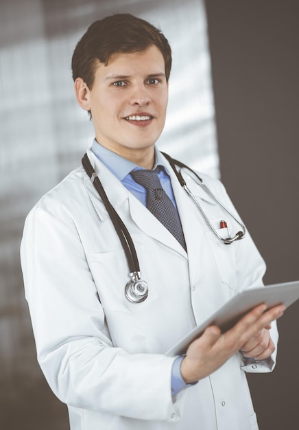 Young friendly doctor is checking some information on his computer tablet. Portrait of a professional physician at work in a clinic. Medicine concept.