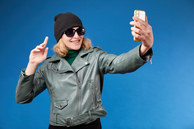 Young friendly fun woman 30s wearing stylish green jacket doing selfie shot on mobile cell phone talking by video call waving hand greeting isolated on blue background studio portrait