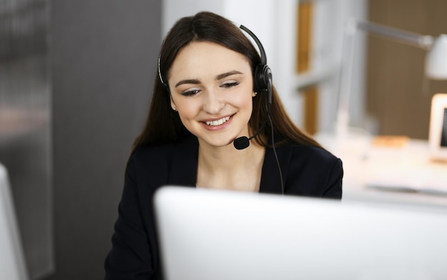 Young friendly girl in headsets is talking to a firm's client, while sitting at the desk in a modern office together with her colleague. Call center operators at work.