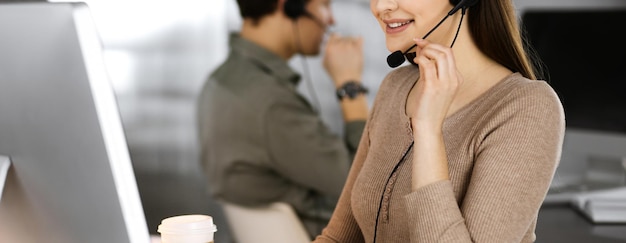 Photo young friendly girl in headsets is talking to a firm's client, while sitting at the desk in a modern office together with her colleague. call center operators at work.