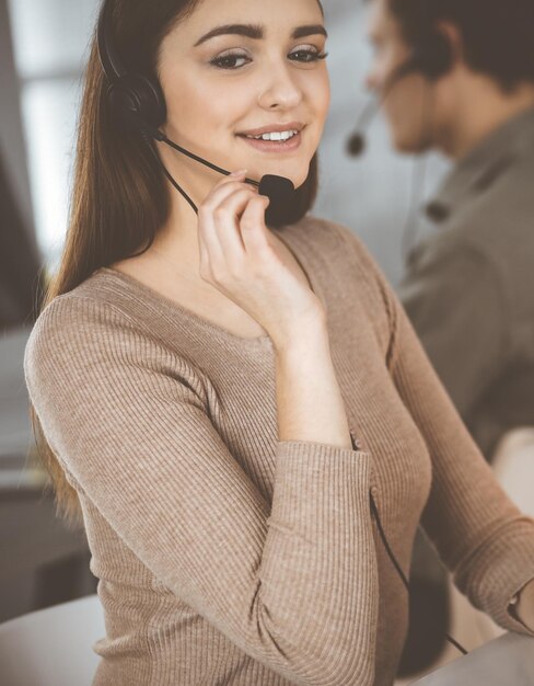 Young friendly girl in headsets is talking to a firm's client, while sitting at the desk in a modern office together with her colleague. Call center operators at work.