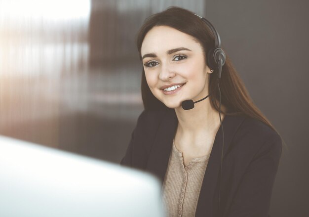 ヘッドセットの若いフレンドリーな女の子は、日当たりの良いオフィスの机に座っている間、会社のクライアントと話しています。仕事中のコール センター オペレーター。