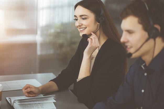 Young friendly girl in headsets is talking to a firm's client, while sitting at the desk in sunny office. Call center operators at work.