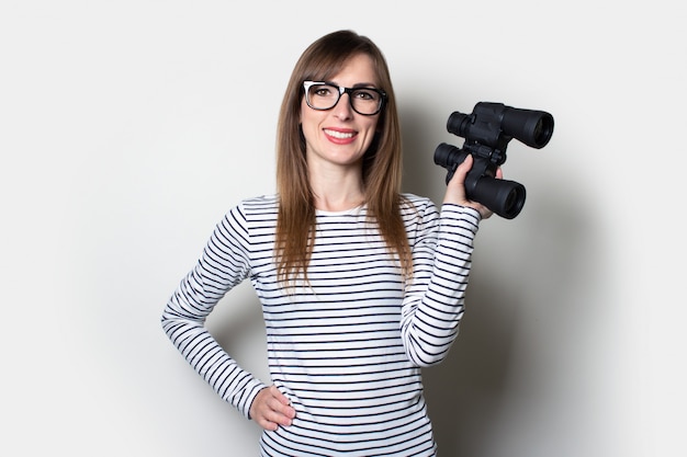 Young friendly girl in a T-shirt holds binoculars on a light space.