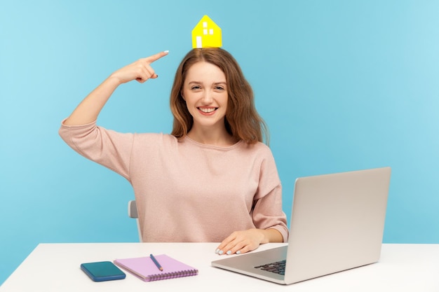 Young friendly happy woman, real estate agent sitting at workplace with laptop and pointing at small paper house on her head, advertising rental service, home purchase. indoor studio shot, isolated