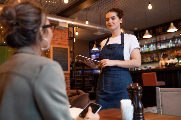 Young friendly waitress with touchpad standing by table with one of clients and taking her order on background of bar counter in restaurant