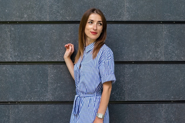 Young friendly woman in a dress looks to the side against the background of a stone wall.