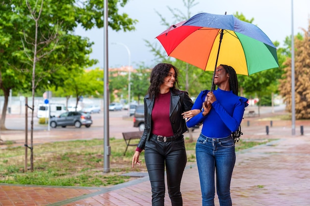 Young Friends Arm in Arm Delighted and Cheerful Strolling in Rainy City with Multicolored Umbrella