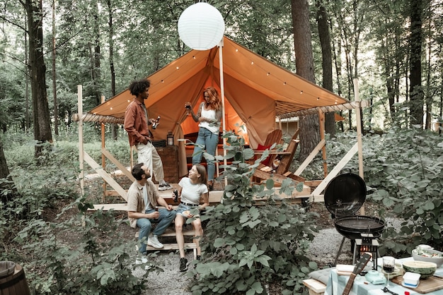 Young friends enjoy sunny evening in forest glamping. People laugh toast clinking beer cider bottles and wine glasses by bonfire near tent. Camping hanging out together, social gathering outdoors