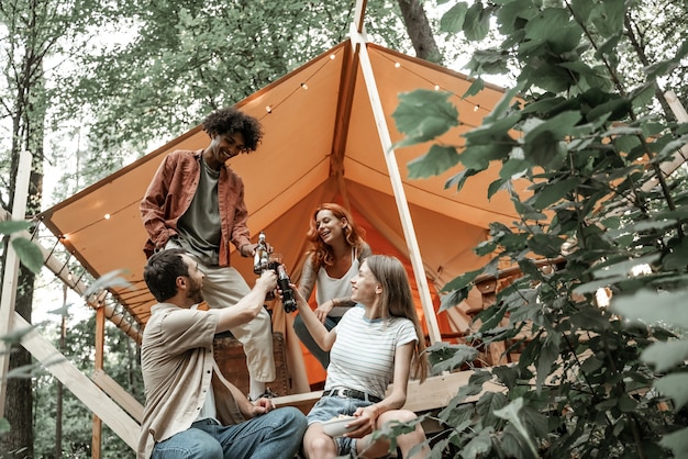 Young friends enjoy sunny evening in forest glamping. people
laugh and toast with beer cider bottles and wine glasses by the
bonfire near tent. camping hanging out together, social gathering
outdoors