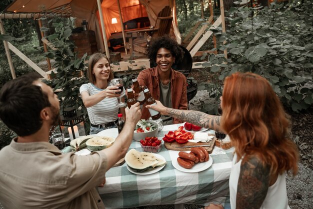 Young friends having dinner at glamping clincking glasses after sunset. Happy millennial gang camping at open air picnic under bulb lights. Spending time with friends outdoors, barbeque party