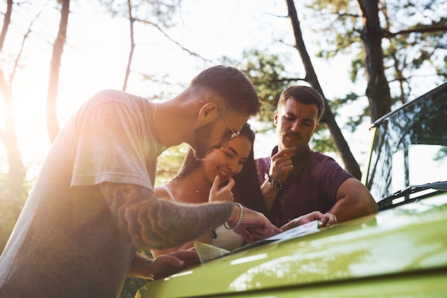 Young friends reading map that is on the hood of modern green jeep in the forest.