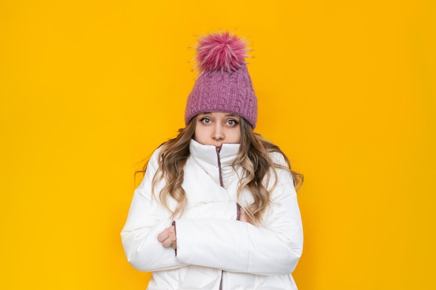 A young frozen blonde woman in a white warm jacket and pink knitted hat shivers hugging herself