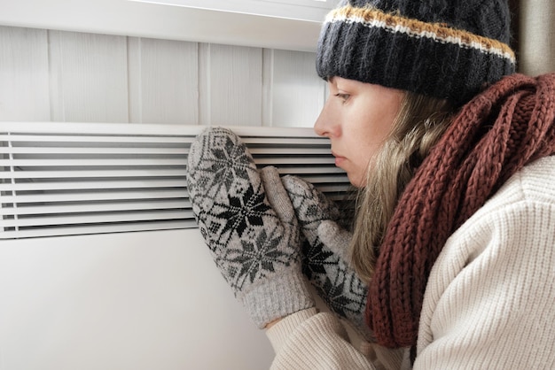 Foto giovane donna congelata che indossa un maglione che trema e si congela per il freddo invernale con un'espressione shock sul viso la ragazza si sta riscaldando le mani con il respiro sul riscaldatore elettrico disagio trascorrendo del tempo a casa