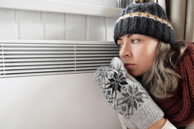 Young frozen woman wearing a sweater shaking and freezing for
winter cold with shock expression on face girl is warming up hands
with breath over electric heater discomfort spending time at
home