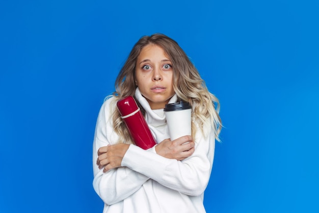 A young frozen woman  in a white wool warm sweater holds a thermos and paper cup in her hands