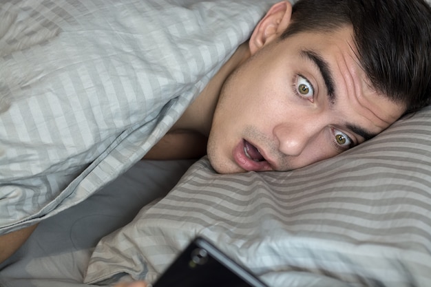 Young frustrated and stressed man is late. He is waking up, looking at smartphone and is shocked.