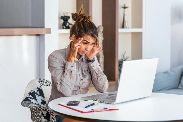 Young frustrated woman working at office desk in front of laptop suffering from chronic daily headaches treatment online appointing to a medical consultation electromagnetic radiation sick pay