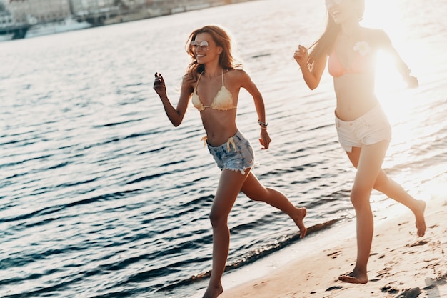 젊고 에너지가 넘칩니다. 해변에서 실행하는 동안 미소 수영복에 두 매력적인 젊은 여성