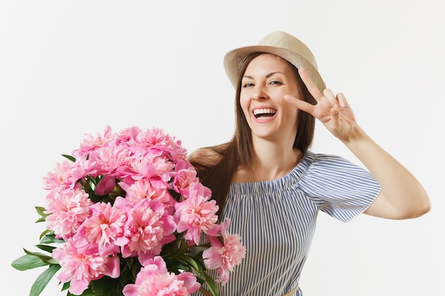 Young fun woman in blue dress, hat holding bouquet of pink peonies flowers, showing victory sign at eye isolated on white background. St. Valentine's Day, International Women's Day holiday concept.