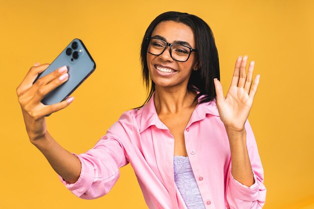 Young funny african american black casual woman grimacing and making selfie on smartphone isolated over yellow background