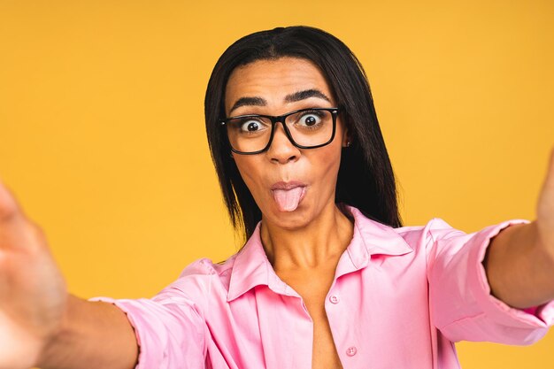 Young funny african american black casual woman grimacing and making selfie on smartphone isolated over yellow background