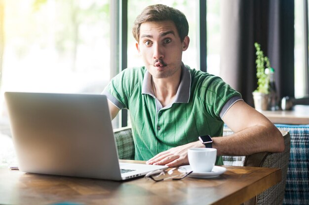 Young funny businessman in green tshirt sitting and working on laptop and looking at camera with fish lips gesture crazy face business and freelancing concept indoor shot near big window at daytime