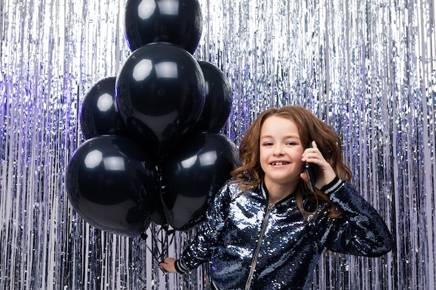 . young funny caucasian girl with helium black balloons in hand communicates on the phone on a shiny tinsel wall