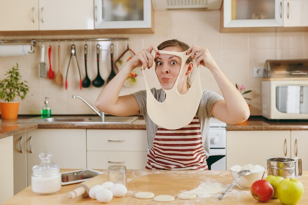 The young funny cheerful and smiling woman puts on a dough with holes on her face and has fun in the kitchen. Cooking home. Prepare food.