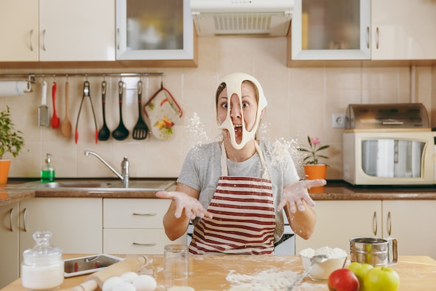 La giovane simpatica donna allegra e sorridente mette su un impasto con i buchi sul viso e si diverte in cucina. cucinare a casa. prepara da mangiare.