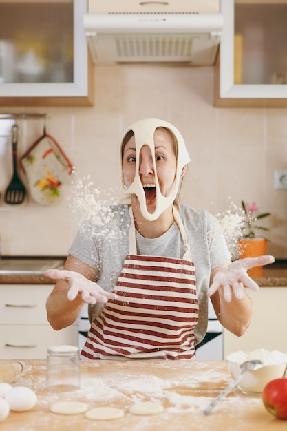 The young funny cheerful and smiling woman puts on a dough with holes on her face and has fun in the kitchen. Cooking home. Prepare food.