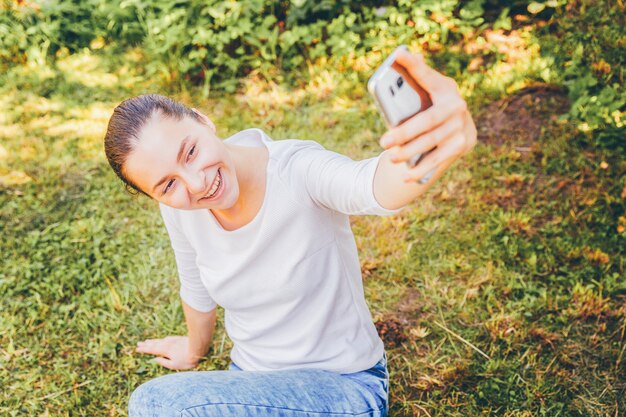 面白い少女は、緑の芝生の公園や庭に座って携帯電話で手からselfieを取る。夏の日にスマートフォンでselfie写真を作る若い魅力的な女性の肖像画。