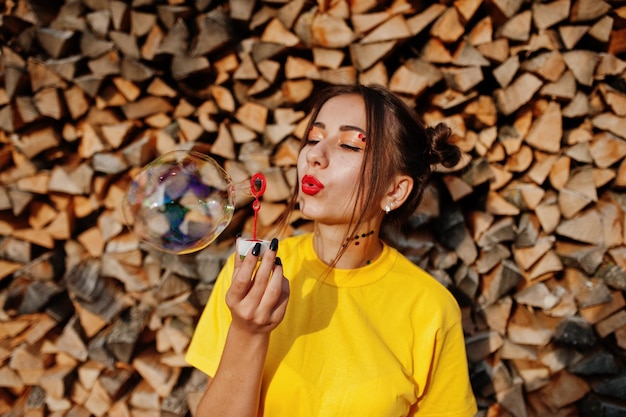 Young funny girl with bright makeup wear on yellow shirt blown soap bubbles against wooden background