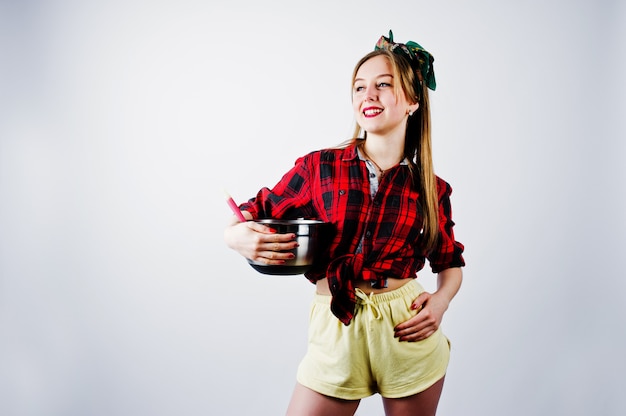 Young funny housewife in checkered shirt and yellow shorts pin up style with saucepan and kitchen spoon isolated