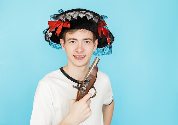 Молодой смешной мальчик-подросток в белой футболке на синем фоне в пиратской шляпе и с пистолетом в руке