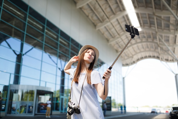 空港で一脚の利己的な棒で携帯電話でselfieをしているレトロなビンテージ写真カメラを持つ若い面白いかわいい旅行者の観光客の女性