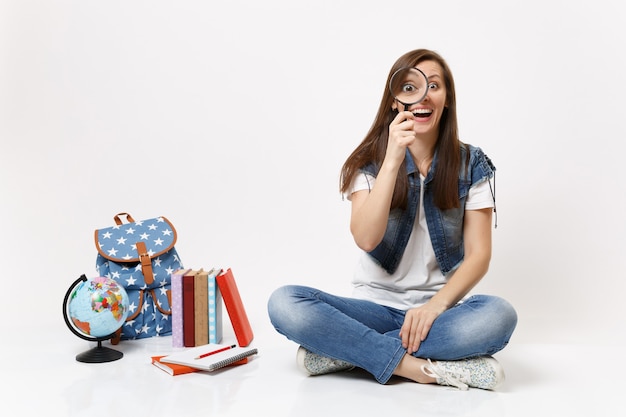 Giovane studentessa carina e divertente che tiene e guarda la lente d'ingrandimento seduta vicino al globo, zaino, libri scolastici isolati