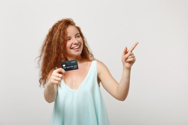 Young funny redhead woman girl in casual light clothes posing isolated on white wall background. People lifestyle concept. Mock up copy space. Holding credit bank card, pointing index finger aside.