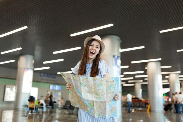 Young funny traveler tourist woman in hat holding paper map, searching route while waiting in lobby hall at international airport