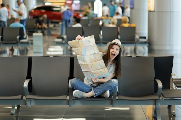 Young funny traveler tourist woman with crossed legs holding paper map, searching route, waiting in lobby hall at international airport