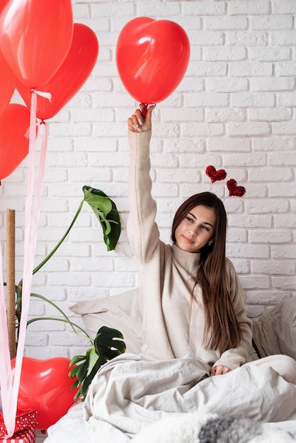 Young funny woman sitting in the bed celebrating valentine day holding balloons