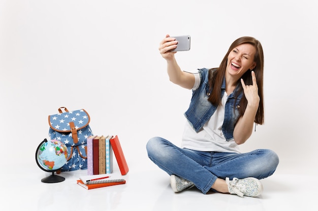 Молодая смешная студентка делает селфи на мобильном телефоне, показывает знак рок-н-ролла, мигает возле школьных учебников с рюкзаком-глобусом